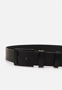 Cinturón de cuero negro con una textura suave, hebilla metálica rectangular y agujeros ajustables. Diseño minimalista con una apariencia elegante.