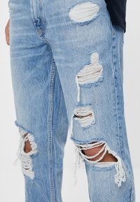 Ljusblå slitna jeans i denim med flera stora revor, fransiga kanter och en avslappnad passform. Rå fåll detalj längst ner.