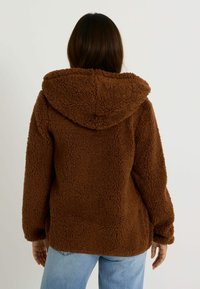Veste à capuche brune, fluffy, avec une texture douce. Présente une coupe ample et des manches longues, avec des détails de couture visibles aux poignets et à l'ourlet.