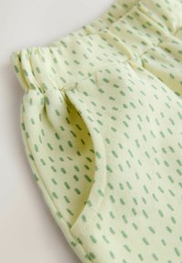 Ljusgul tyg med små gröna prickmönster, utrustad med elastisk midjeresår och sidofickor. Mjuk textur, avslappnad design.