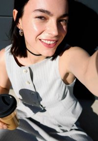 Svetlo šedé bezrukávové šaty s jemným prúžkom, okrúhlym výstrihom a gombíkmi. Osoba drží šálku kávy a má na sebe čierny choker.