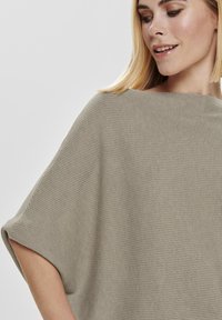 Taupe oversized gebreide poncho met geribbelde textuur, brede mouwen en bootnek. Lichtgewicht en zachte stof. Minimalistisch ontwerp.