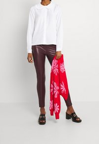 Blusa bianca a maniche lunghe con colletto arricciato, leggings in similpelle marrone e sciarpa rossa con motivi floreali rosa. Sandali neri con tacco a blocco.