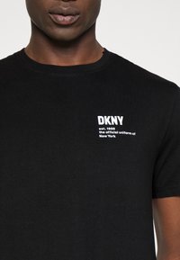 Detailní záběr osoby nosící černé tričko s bílým nápisem "DKNY est. 1989 oficiální uniforma New Yorku" na hrudi.