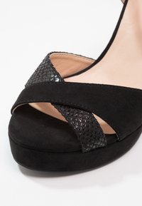 Sandale plateforme noire avec des brides croisées, l'une en daim lisse et l'autre en matériau texturé brillant, présentée sur un fond clair.
