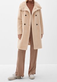 Beige dubbelknäppt kappa i mjukt, texturerat tyg, med stor krage och svarta knappar. Bärs över en brun polotröja och beige byxor.