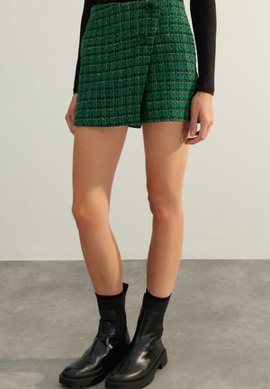 Frau trägt einen grünen Tweed-Minirock mit Knöpfen und schwarze Stiefeletten auf grauem Boden vor einer schlichten hellen Wand.