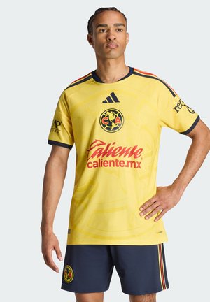 Mand iført en gul Club América fodboldtrøje med rød sponsor tekst og marineblå shorts med klublogo, poserer med den ene hånd på hoften.