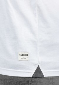 Camiseta blanca de algodón con una etiqueta de marca rectangular en la parte inferior izquierda que dice "SOLID" y "EST. 1999", y un detalle de dobladillo triangular.