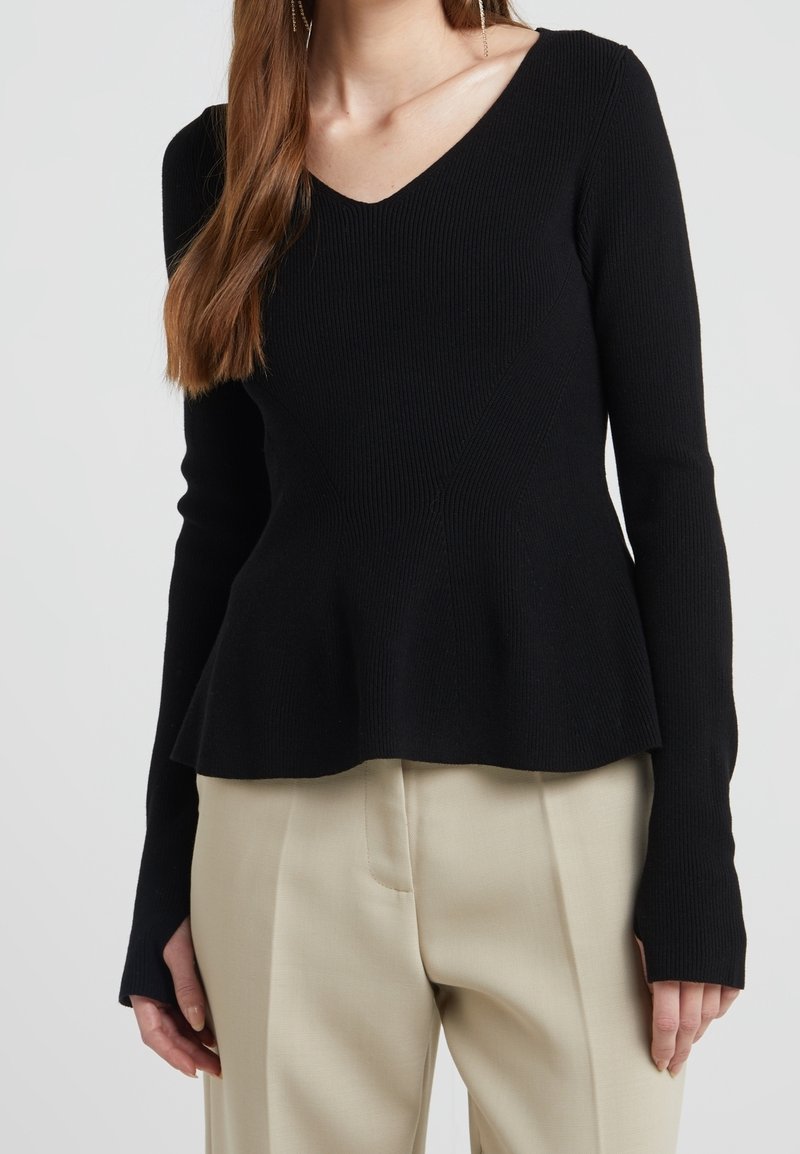 Femme portant un haut noir en tricot côtelé à manches longues avec décolleté en V et ourlet peplum, associé à un pantalon beige taille haute sur un fond neutre.