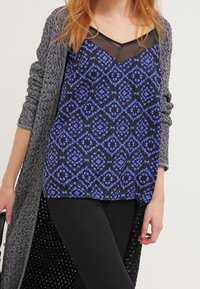 Top negro con patrón geométrico púrpura y escote transparente, combinado con un cárdigan largo de textura gris. Modelo llevando pantalones negros.