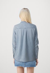 Lichtblauwe spijkerblouse met lange mouwen, knoopmanchetten en een gebogen zoom. Bevat een yoke en subtiele structuur. Draagt samen met een blauwe spijkerrok.