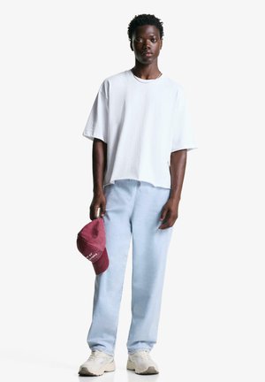 Witte oversized t-shirt, lichtblauwe jeans en crème sneakers. Maroon pet in één hand met witte tekst. Eenvoudige, casual outfit.