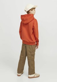 Felpa arancione con cappuccio e vestibilità larga, abbinata a pantaloni cargo color khaki con tasche laterali. Cappellino chiaro e scarpe beige.