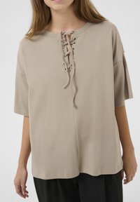 Taupe top met korte mouwen en een vetersluiting aan de voorkant, voorzien van metalen oogjes en een relaxte pasvorm, gemaakt van zachte, gladde stof.
