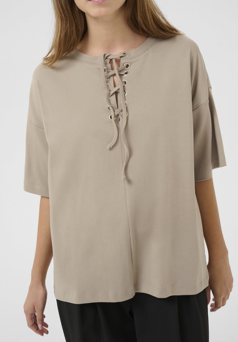 Taupe top met korte mouwen en een vetersluiting aan de voorkant, voorzien van metalen oogjes en een relaxte pasvorm, gemaakt van zachte, gladde stof.