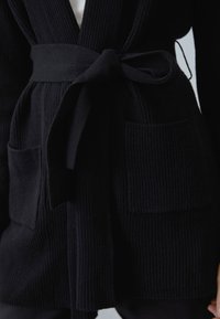 Schwarze Strickjacke mit gerippter Textur, ausgestattet mit einem Gürtel zum Binden in der Taille, zwei vorderen Taschen und einem Schalkragen.