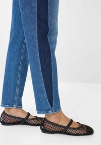 Gambe che indossano jeans blu bicolore abbinati a ballerine Mary Jane in rete nera su un pavimento bianco e uno sfondo semplice.