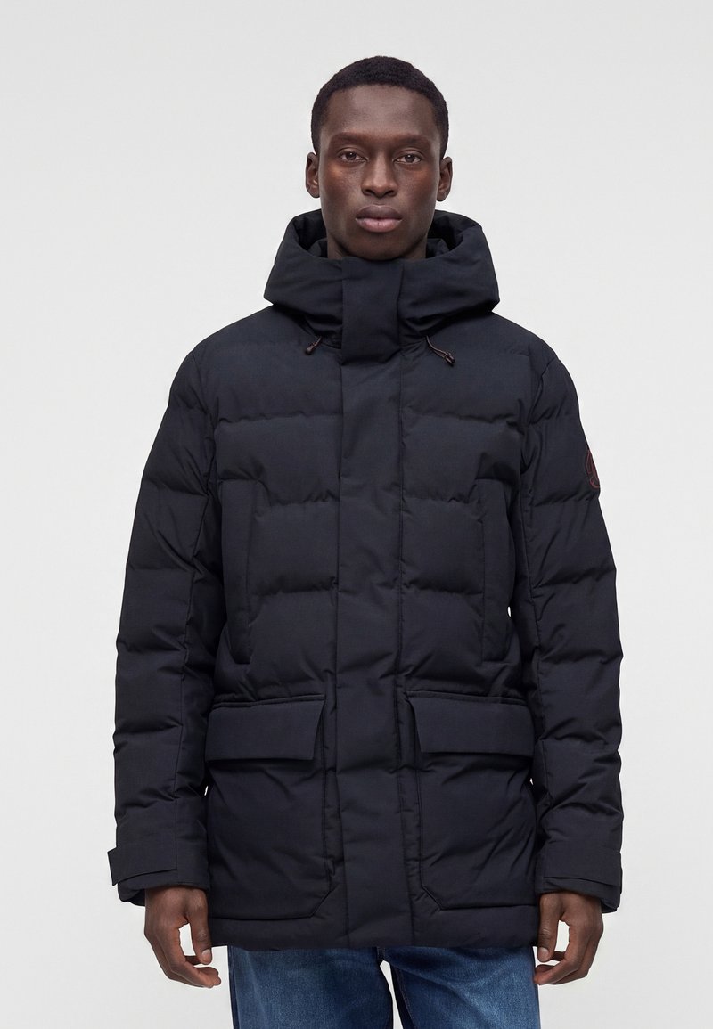 Νέος άνδρας με μαύρο επενδυμένο χειμερινό μπουφάν με ψηλό γιακά και κουκούλα, που στέκεται μπροστά σε ένα μονόχρωμο ανοιχτό γκρι φόντο.