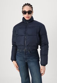 Tengerészkék puffertakaró, rövidített fazonnal, magas gallérral és elasztikus mandzsettákkal. Magas derekú kék farmernadrággal párosítva. Simább, matt textúrával.