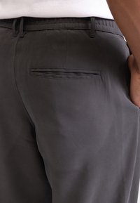Primer plano de una persona que viste pantalones gris oscuro con un bolsillo trasero y una camisa blanca metida por dentro.