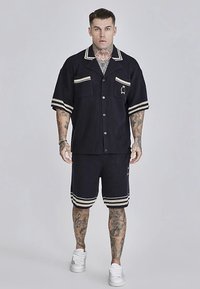 Sötétkék két részes öltözék, texturált anyagból; rövid ujjú, gombos ingből és hozzáillő rövidnadrágból áll, csíkos díszítéssel.