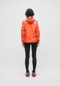 Veste orange imperméable avec capuche, coupe ajustée, accents réfléchissants, associée à des leggings noirs et des chaussures de sport noir-rouge.