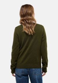Femme aux cheveux châtain clair ondulés porte un pull vert foncé et un jean bleu, vue de dos sur un fond blanc uni.