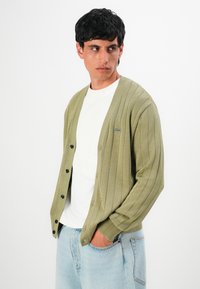 Homem com um cardigan canela canelado sobre uma camisa branca, com a mão no bolso, olhando de lado contra um fundo branco.