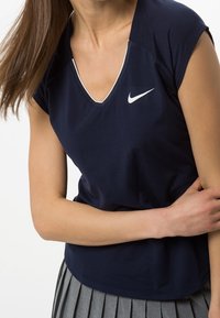 Kvinna iklädd en marinblå Nike V-ringad kortärmad topp och grå plisserad kjol, som håller sin vänstra arm med sin högra hand.