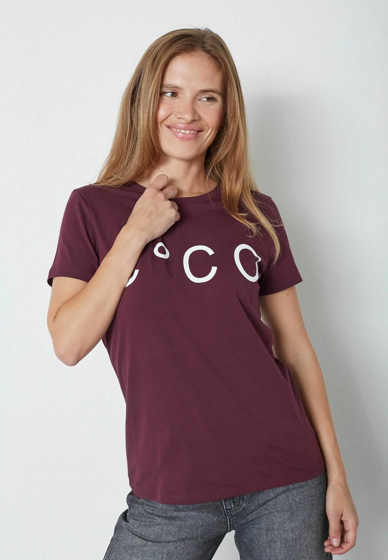 Burgundin t-paita, joka on valmistettu puuvillasta, lyhythihainen ja siinä on valkoista tekstiä edessä. Yhdistetty harmaisiin, slim fit -farkkuihin. Sujuva pinta.