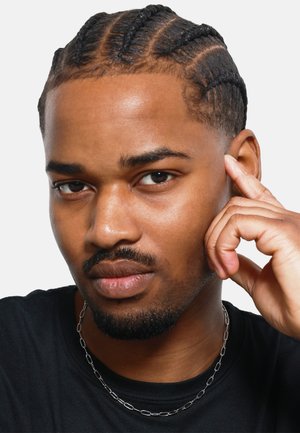 Homme avec des cheveux tressés portant un T-shirt noir et un collier en chaîne argenté. Il a une barbe courte et touche son visage avec une main.