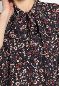 Gros plan sur un blouse floral sombre avec des fleurs rouges, blanches et bleues et un nœud noué au niveau du col.