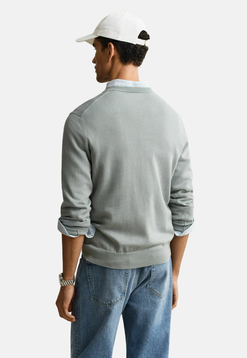 Hombre vistiendo un suéter gris claro sobre una camisa a rayas con las mangas remangadas, jeans azul claro, gorra blanca y reloj de pulsera plateado, de espaldas.