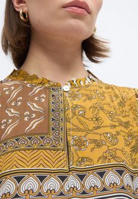 Κοντινό πλάνο ενός ατόμου που φοράει μπλούζα με κίτρινο και καφέ σχέδιο, με λουλουδένια και γεωμετρικά μοτίβα, με ορατά χρυσά σκουλαρίκια κρίκους.