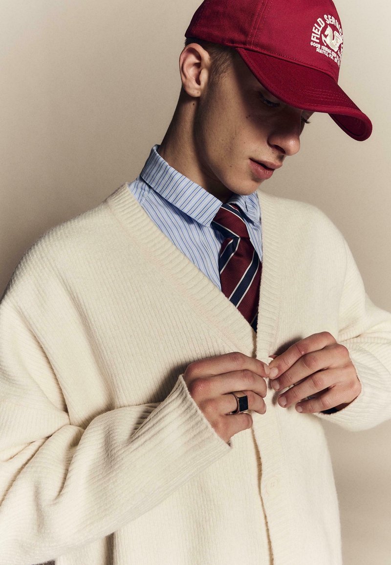 Jeune homme boutonnant un cardigan en laine crème, portant une casquette rouge, une chemise bleue rayée et une cravate rayée rouge sur un fond uni.