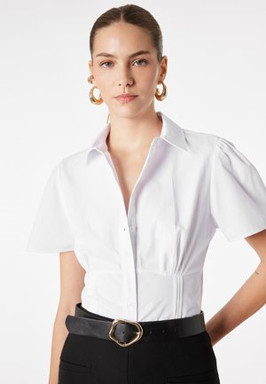 Femme aux cheveux tirés en arrière, portant des boucles d’oreilles créoles dorées, un chemisier blanc cintré à manches courtes, un pantalon noir et une ceinture noire avec une boucle dorée.