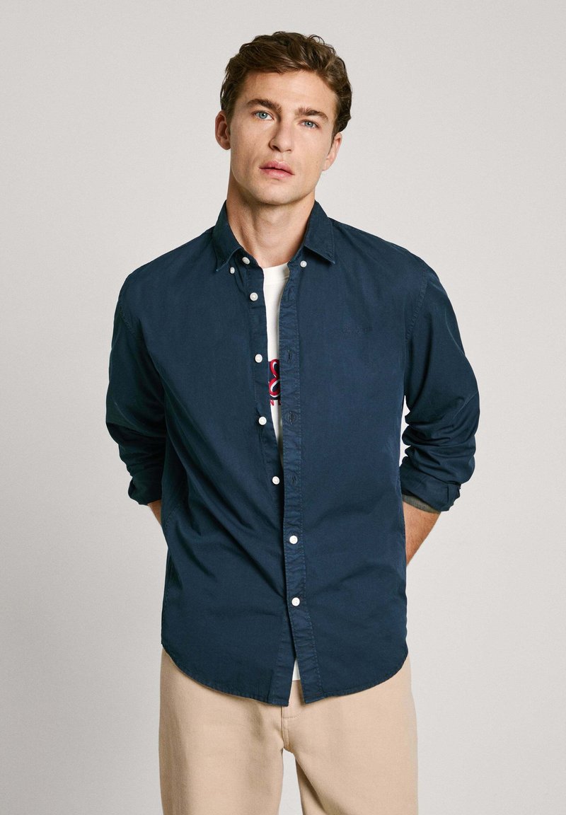 Camisa de botones azul marino con una textura suave, con mangas largas arremangadas en los puños, llevada sobre una camiseta gráfica blanca.