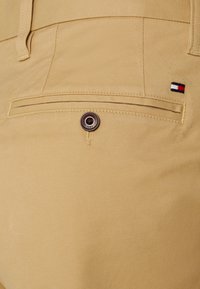 Khaki spodnie o gładkiej fakturze, z tylną kieszenią z srebrnym guzikiem i małym detalem logo w kolorach czerwonym, białym i niebieskim.