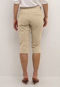 Beige capribyxor med en figurnära design, tillverkade av mjuk tyg, med två bakfickor och en fåll som slutar nedanför knäet. Bärs med sandaler.