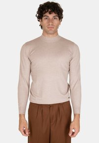 Maglione beige chiaro a maniche lunghe con alto colletto, vestibilità aderente e texture morbida, abbinato a pantaloni marroni a pieghe.