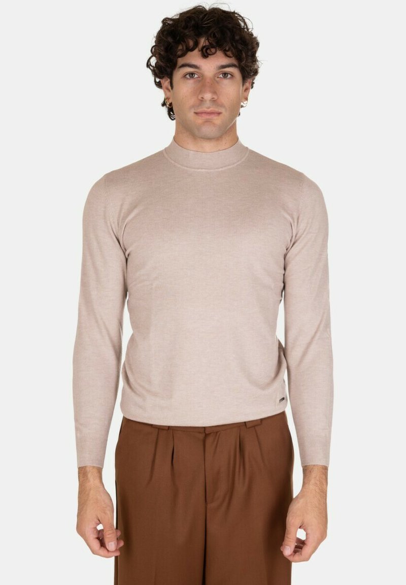 Maglione beige chiaro a maniche lunghe con alto colletto, vestibilità aderente e texture morbida, abbinato a pantaloni marroni a pieghe.