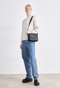 Bolso de hombro de cuero negro con forma rectangular, que cuenta con una correa larga y un sutil logotipo. El modelo lleva una sudadera gris y jeans azules.
