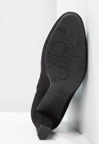 Černá semišová lodička na vysokém podpatku s špičatou špičkou. Podešev zdobená raženými vzory a logem, ukazující texturu a plochý design podpatku.