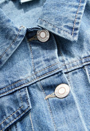 Világoskék farmerdzseki, amely galléros nyakkivágással, két elülső zsebbel, fém patent gombokkal és kontrasztos öltési részletekkel rendelkezik.