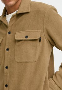 Beige fleeceshirt-jack met een knoopsluiting, twee borstzakken en zwarte knopen. Zachte textuur met een casual pasvorm en een standaard kraag.