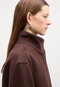 Hnědý kabát s vysokým límcem a měkkou strukturou, s strukturovanými švy a hladkým kovovým kruhovým náušnicí na pravém uchu modelu.