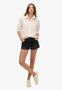 Biała lniana koszula z guzikami, czarne szorty dżinsowe z postrzępionymi brzegami i kremowe sneakersy; model stoi z dłońmi w kieszeniach na jednolitym tle.