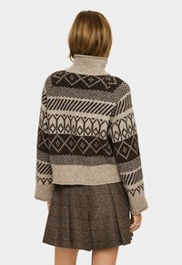 Person in einem braun gemusterten Rollkragenpullover und einem braunen Faltenrock, mit dem Rücken zur Kamera und sichtbarem hellbraunem Haar.