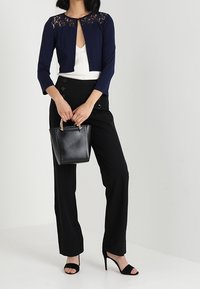 Marinblå kort cardigan med spetsdetaljer vid halsringningen, ihop med högmidjade svarta byxor och en svart handväska. Svarta klacksandaler.